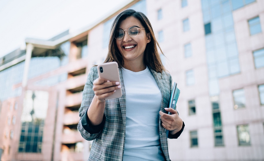 blog_-_how_the_new_whatsapp_interactive_button_helps_ace_conversational_automation A young woman is engrossed in her phone, browsing through an interactive Whatsapp message template while standing in front of a building.