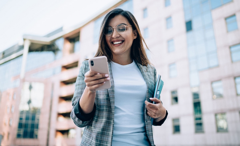 A young woman is engrossed in her phone, browsing through an interactive Whatsapp message template while standing in front of a building.