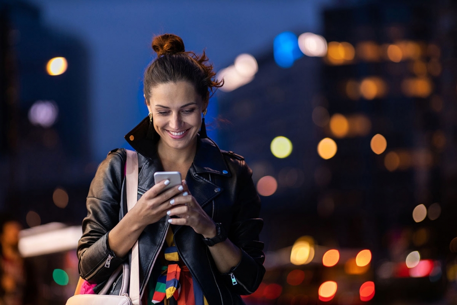 A woman is looking at her phone in a city at night, possibly browsing through WhatsApp notifications after opting in to receive them.