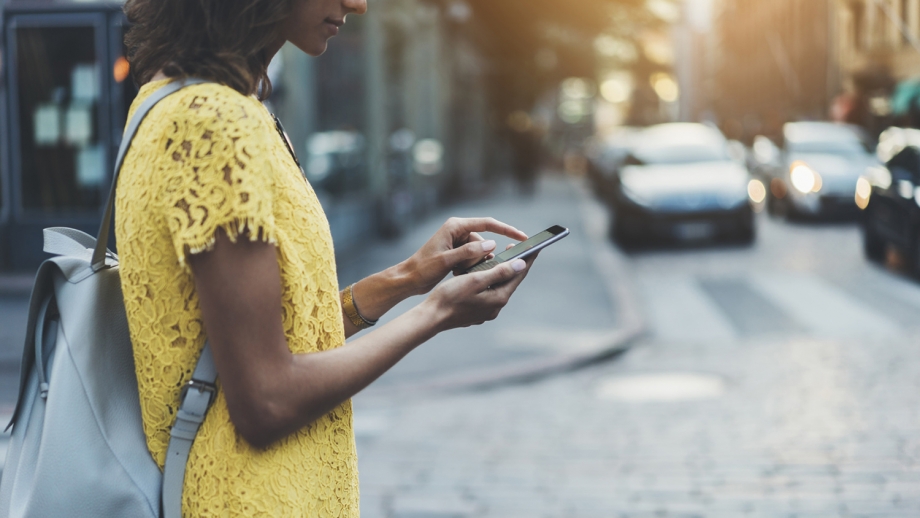 A woman in a yellow dress is using her cell phone on the street, highlighting the vulnerability of SMS-Based Authentication.