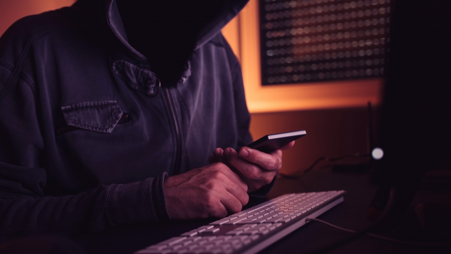 A person in a hoodie is using a cell phone in front of a computer.