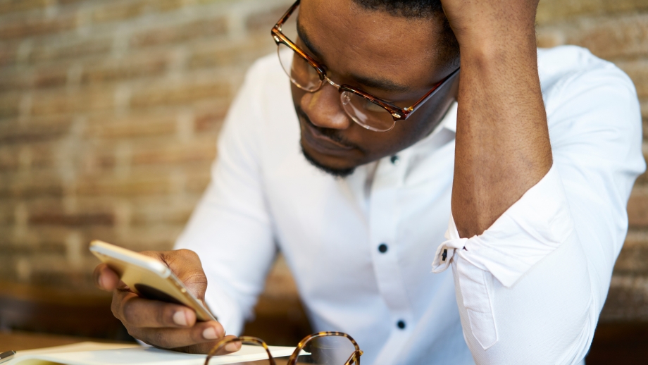 A man with glasses is looking at his phone while sitting at a table, potentially worried about his WhatsApp Business account being banned.