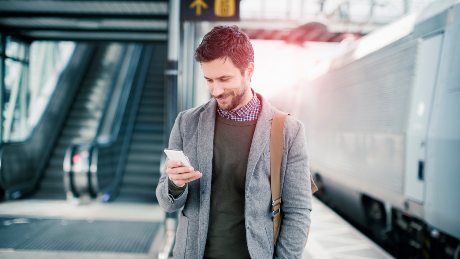 A man is standing near a train station, engrossed in his phone.