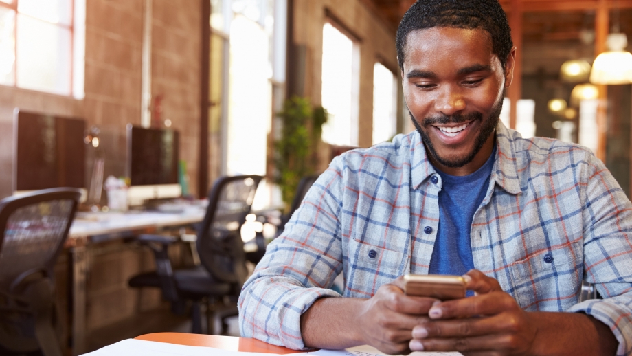 A smiling black man using a cell phone in an office while engaged in eCommerce activities.