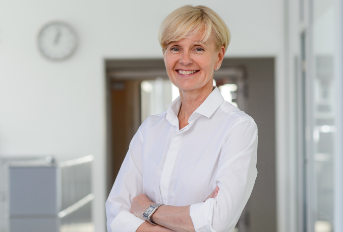 A smiling business woman in a white shirt standing in an office, showcasing the About Us section.